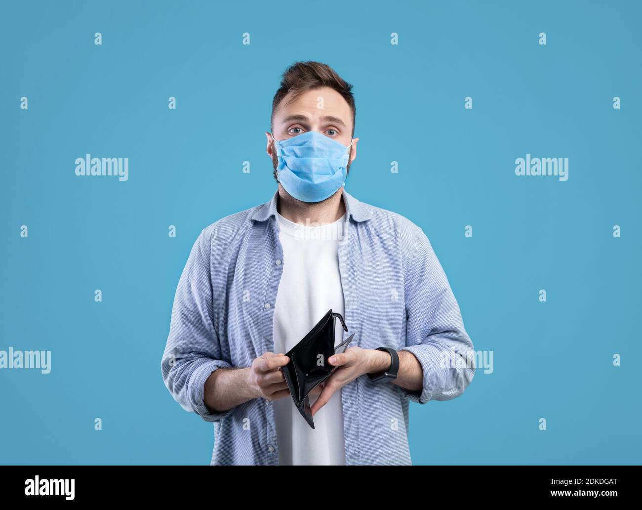 Crisi economica del coronavirus. Ragazzo giovane in maschera medica mostrando portafoglio vuoto su sfondo blu studio Foto Stock