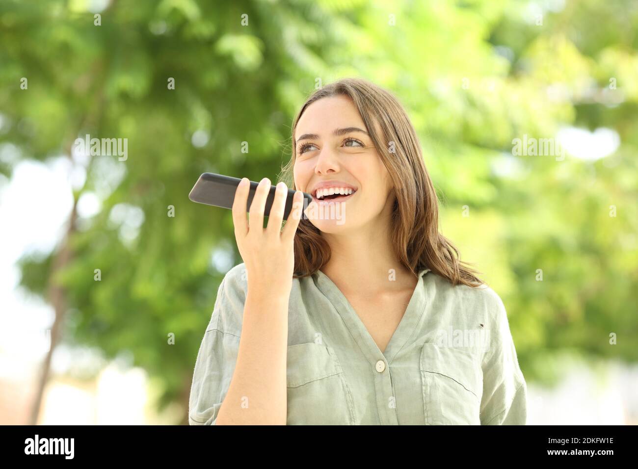 Donna felice che registra un messaggio audio sullo smartphone che cammina dentro un parco Foto Stock