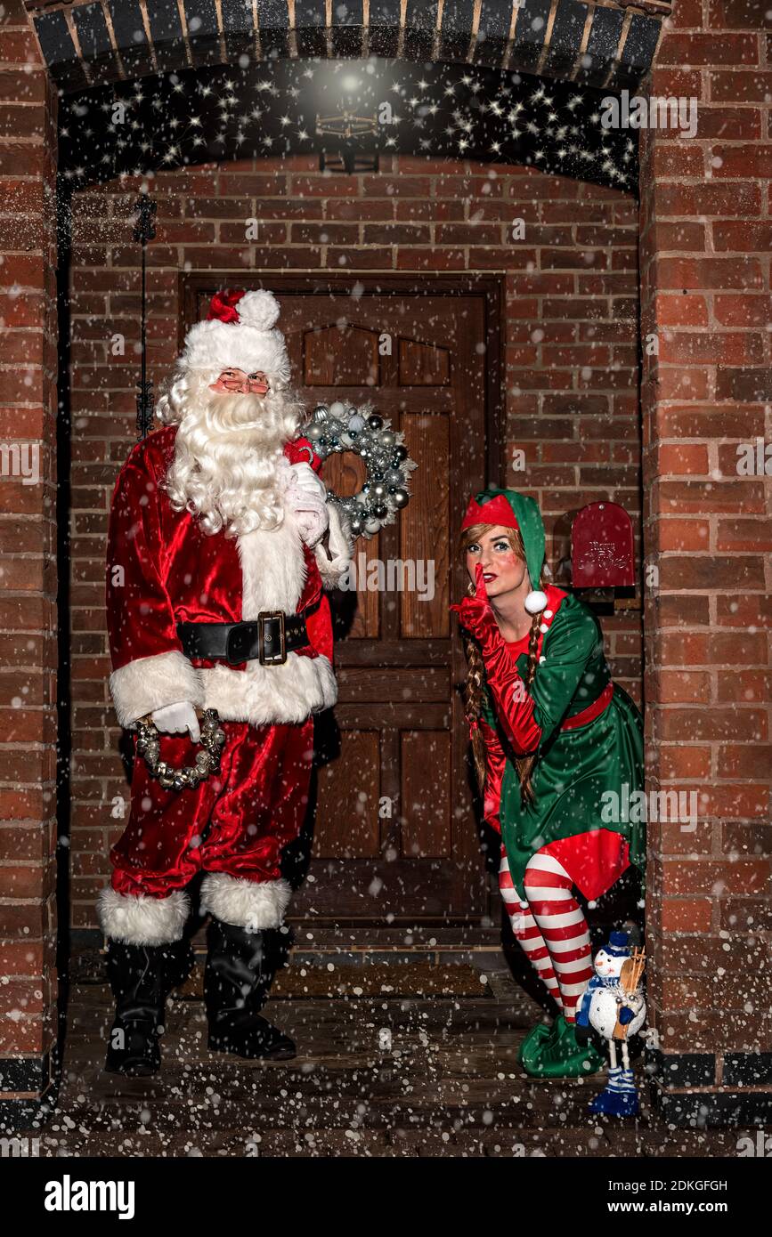 Regno Unito, Quorn - Dicembre 2020: Santa e un Elf si sbirciano fino a una porta d'ingresso, nella neve, mentre consegnano i regali Foto Stock