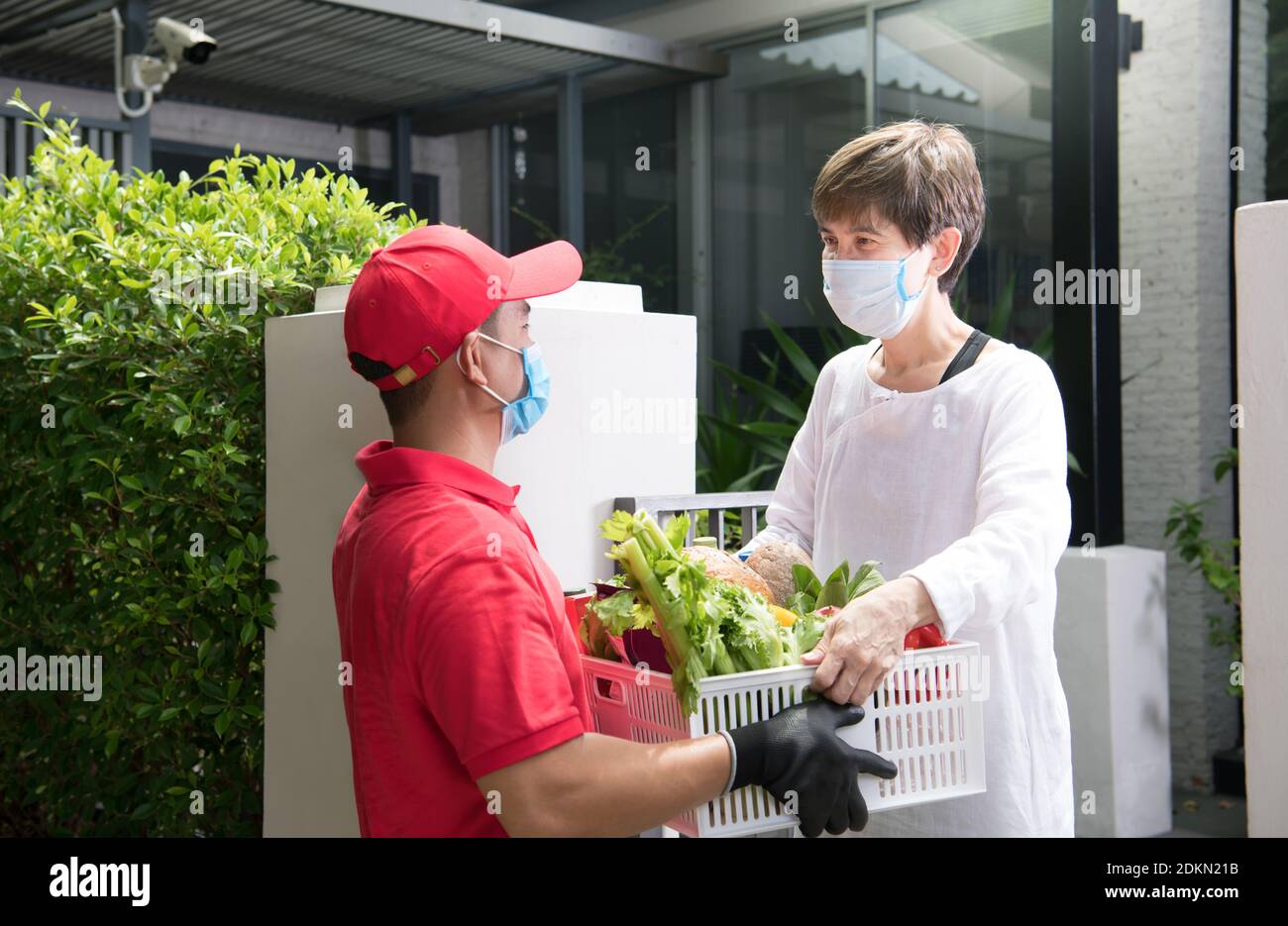 Parto asiatico uomo che indossa maschera e guanti in uniforme rossa che consegna drogheria scatola di cibo, frutta, verdura e bevande al destinatario durante COVID Foto Stock