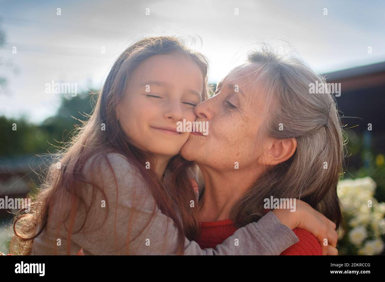 La nonna anziana con i capelli grigi che indossa il maglione rosso con la sua nipote piccola si aggingono nel giardino e durante la giornata di sole all'aperto, la madre Foto Stock