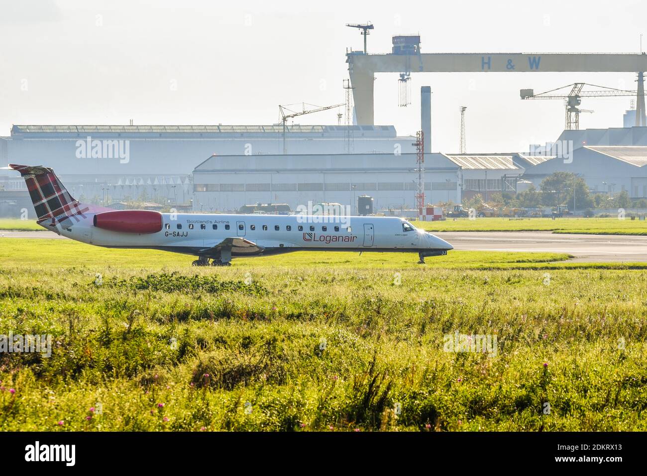 Loganair Embraer EMB-145 G-SAJJ in partenza dall'aeroporto BHD di Belfast City, Irlanda del Nord con l'iconica gru a portale gigante Sampson del cantiere navale di Belfast Foto Stock