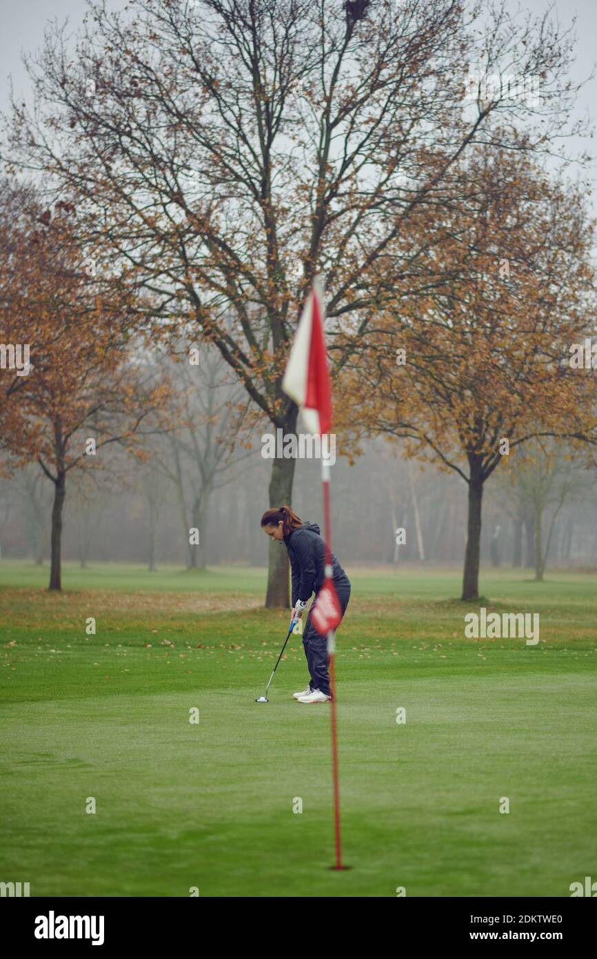 Medio-invecchiato golfer della donna di misura che mette per il buco sul verde visto dalla bandiera che si concentra per l'oscillazione successiva Foto Stock
