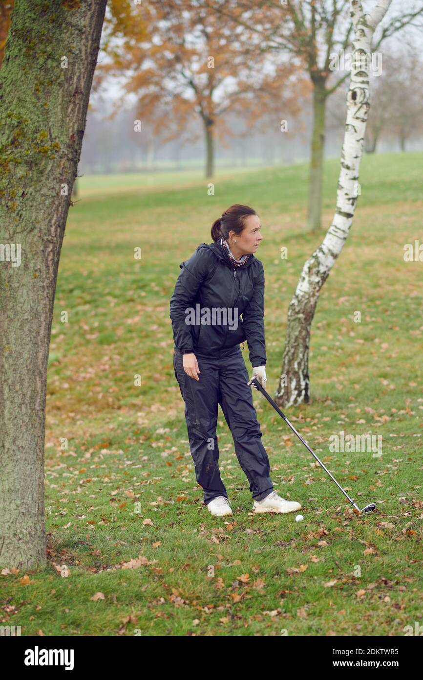 Donna golfer che gioca un tondo di golf in piedi tra gli alberi in una fredda giornata invernale con club in mano staring oltre il fairway Foto Stock