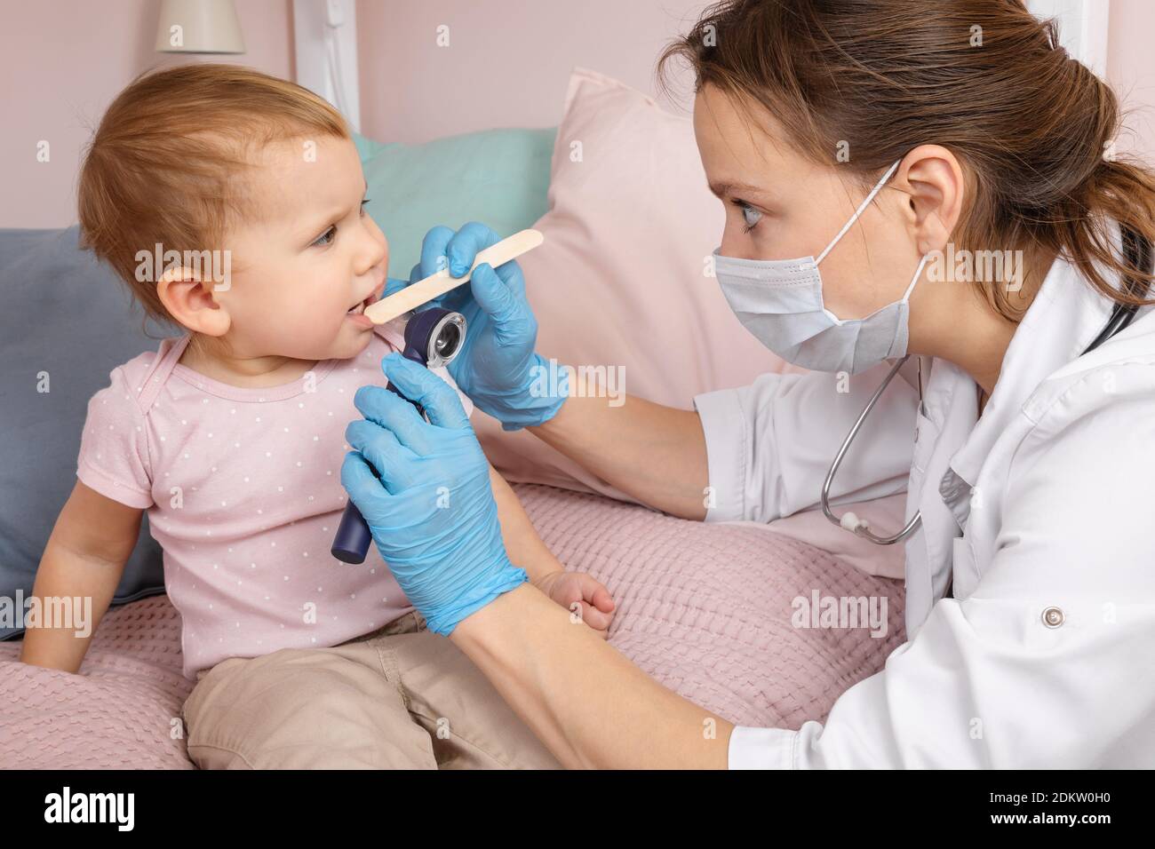 Pediatra esamina la gola irritata di una bambina a casa durante la quarantena del coronavirus COVID-19. Medico che usa il depressor della lingua di legno e la torcia Foto Stock