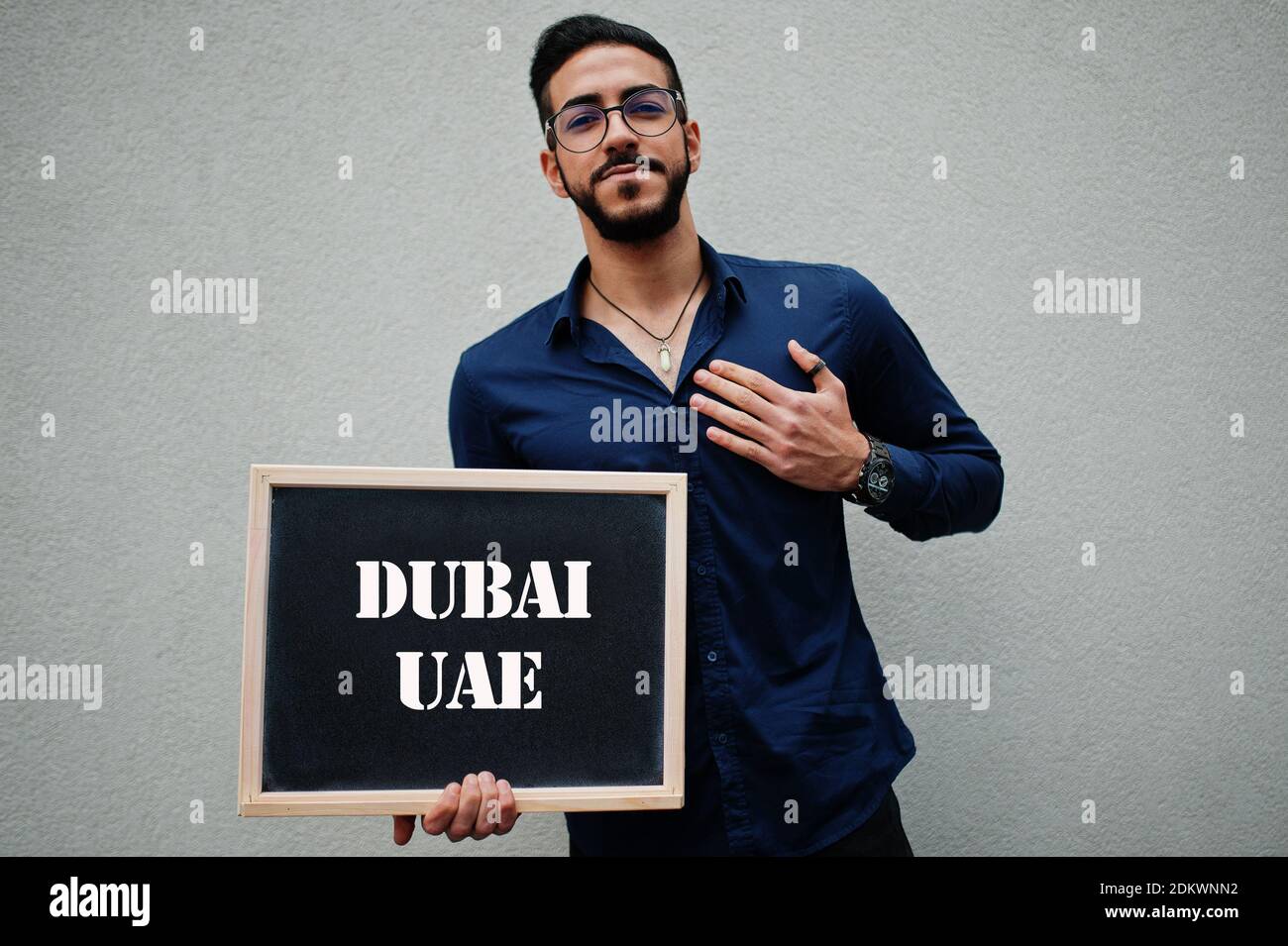 Uomo arabo indossare camicia blu e occhiali tenere tavola con iscrizione Dubai UAE. Le città più grandi del mondo islamico concetto. Foto Stock