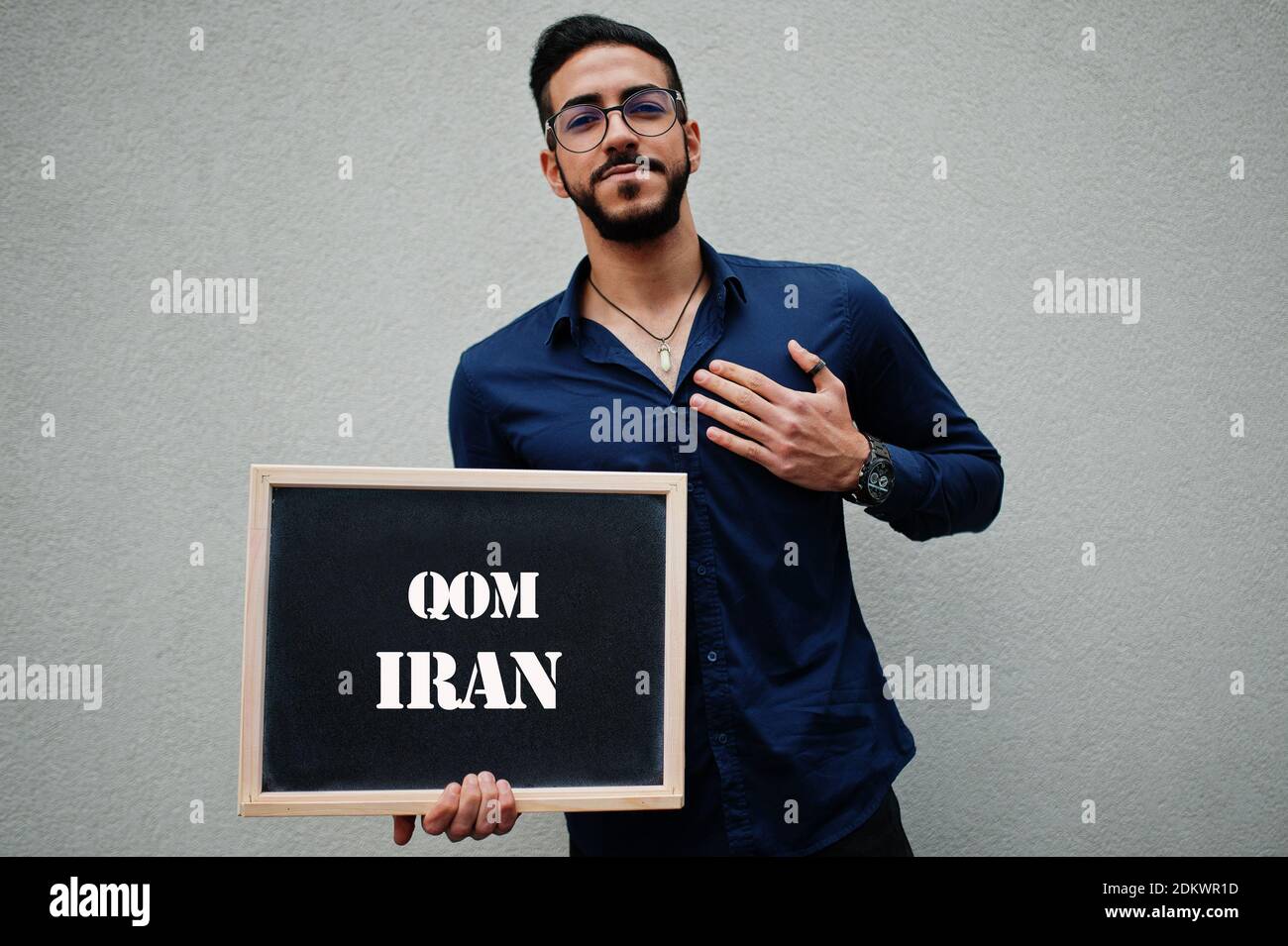 Uomo arabo indossare camicia blu e occhiali tenere tavola con iscrizione Qom Iran. Le città più grandi del mondo islamico concetto. Foto Stock