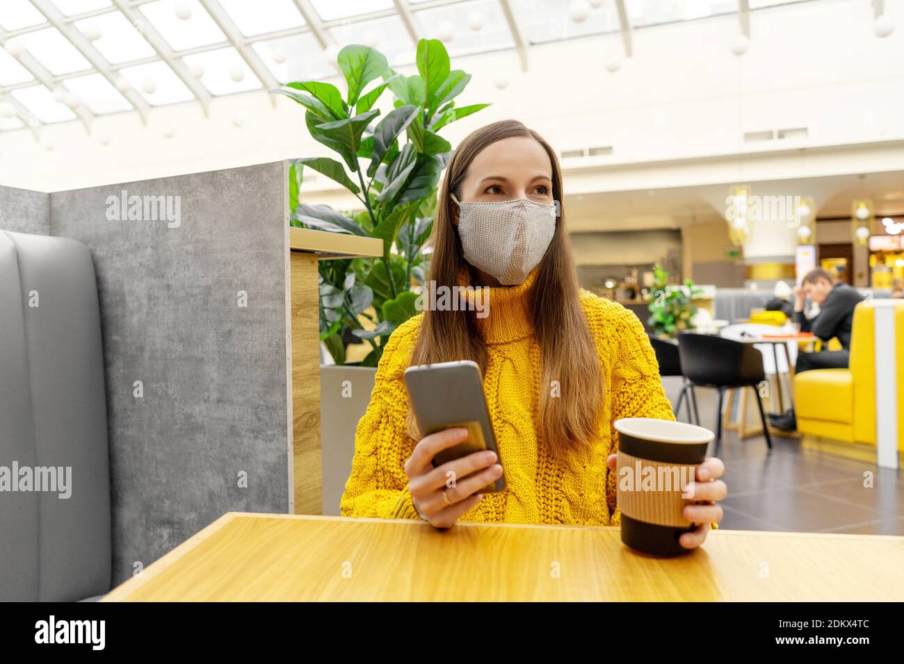 Bruna giovane donna felice in maschera protettiva con tazza di caffè seduto al tavolo in centro commerciale o caffè. Colori giallo e grigio. Foto Stock