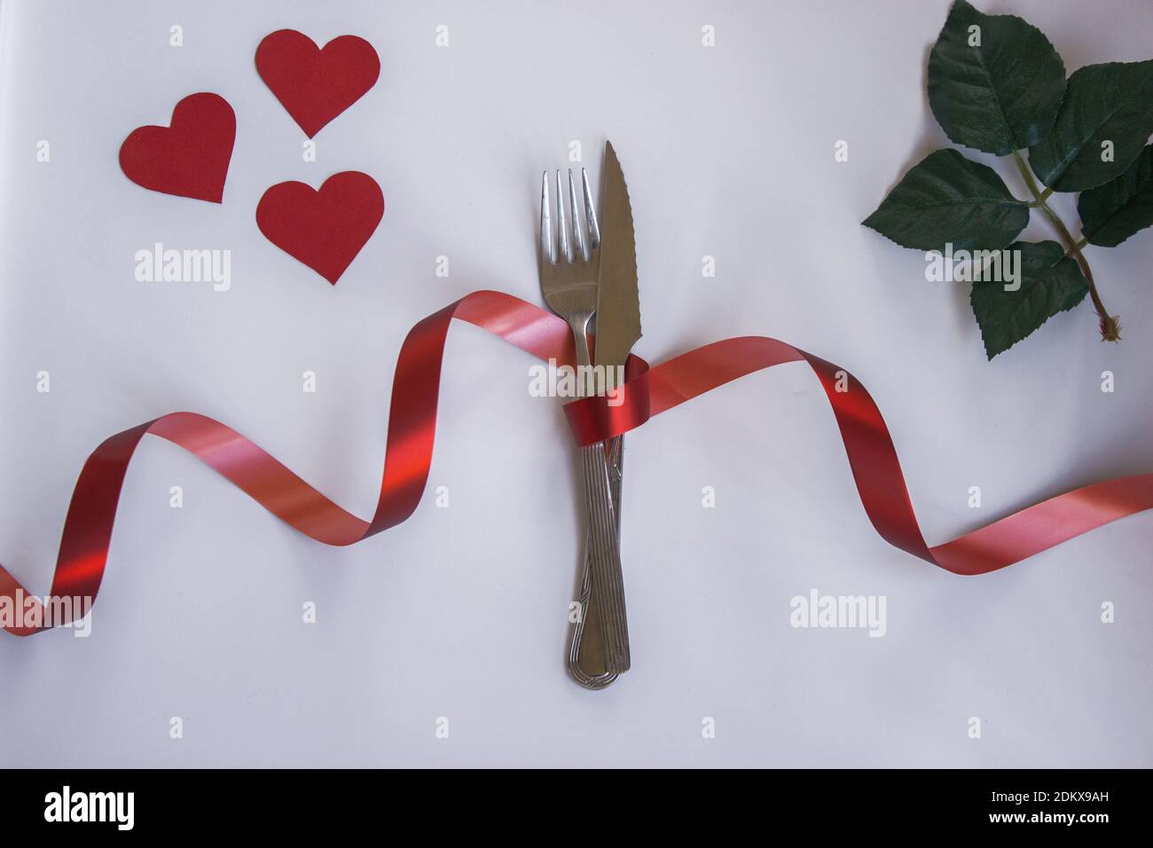 Forcella e coltello argentati legati con un filo rosso. Su uno sfondo bianco isolato e cuori rossi come simbolo del giorno di San Valentino. Concetto di San Valentino 2 Foto Stock