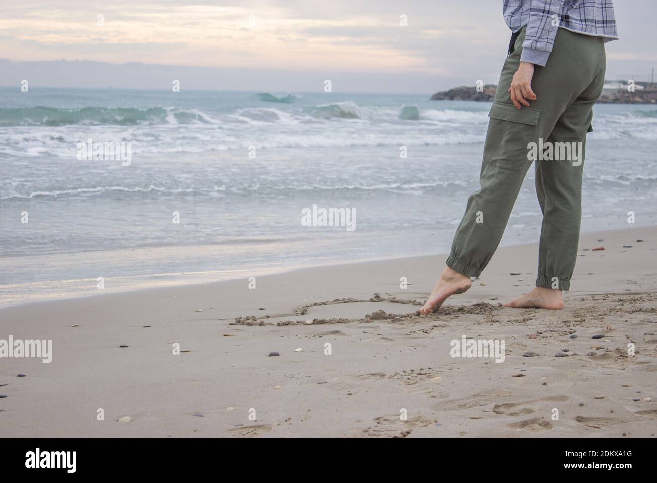 Immagine concettuale orizzontale con sfondo oceano. Una donna irriconoscibile attira un cuore sulla sabbia con il piede il 14 febbraio. San Valentino Foto Stock