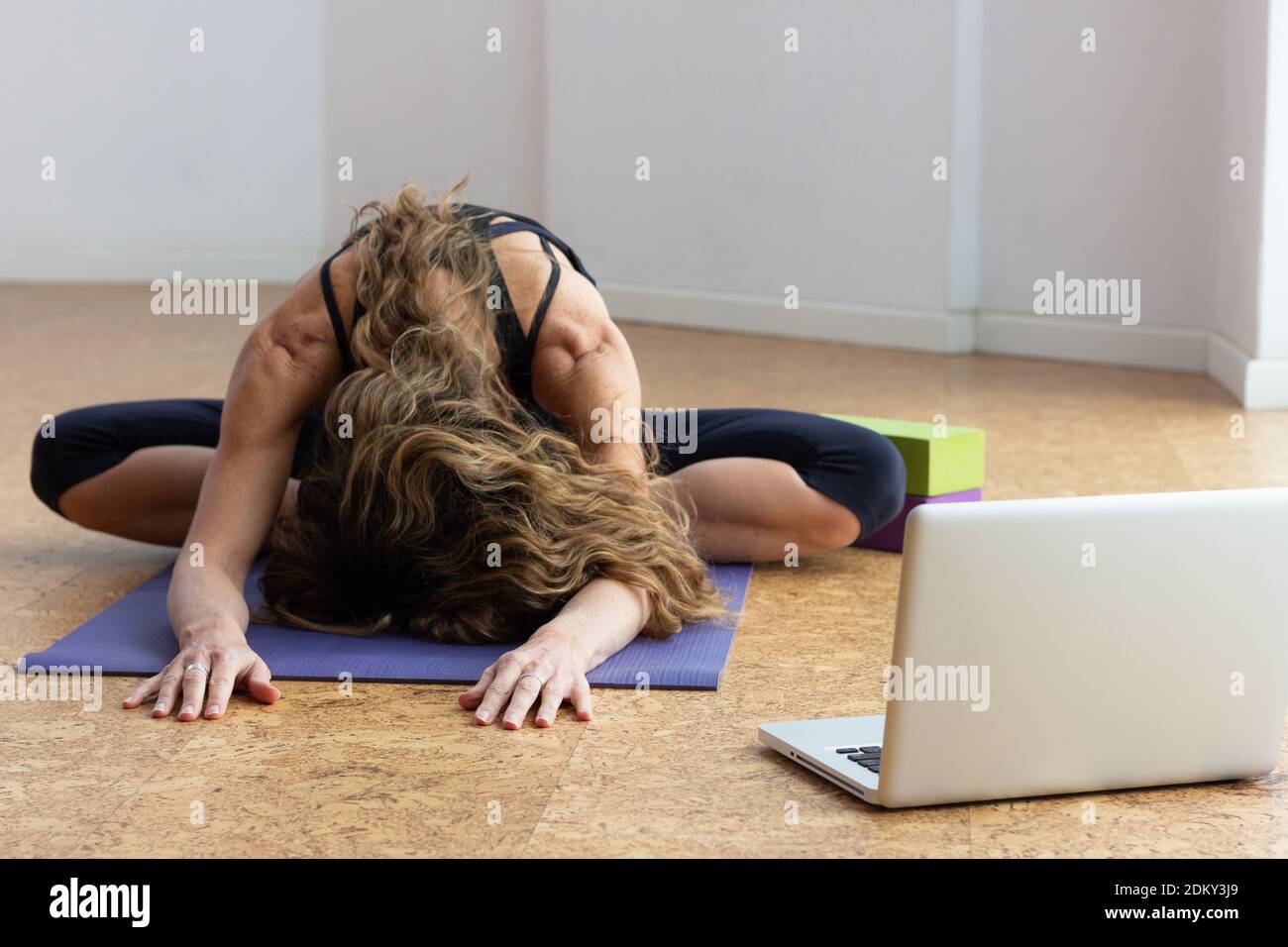Donna flessibile seduta in posizione farfalla con testa giù sul tappetino yoga e laptop sul lato. Yogi femmina che fa la classe in linea all'interno. Concetto di blocco Foto Stock