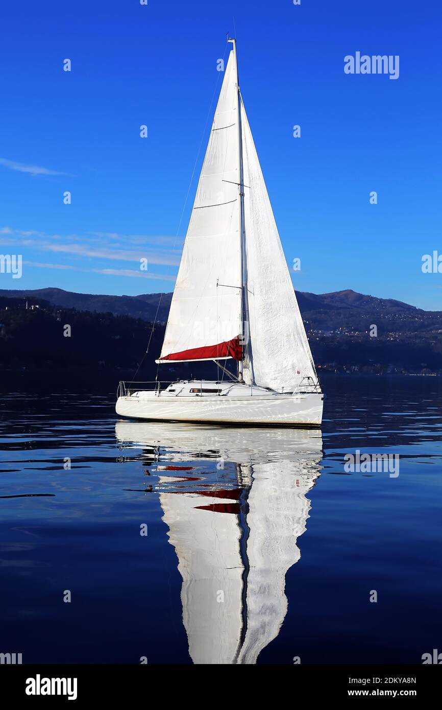 Barca a vela bianca che galleggia sulle acque blu profonde del lago, mare con riflessi sullo sfondo delle montagne vicino all'isola di San Giulio, Italia Foto Stock