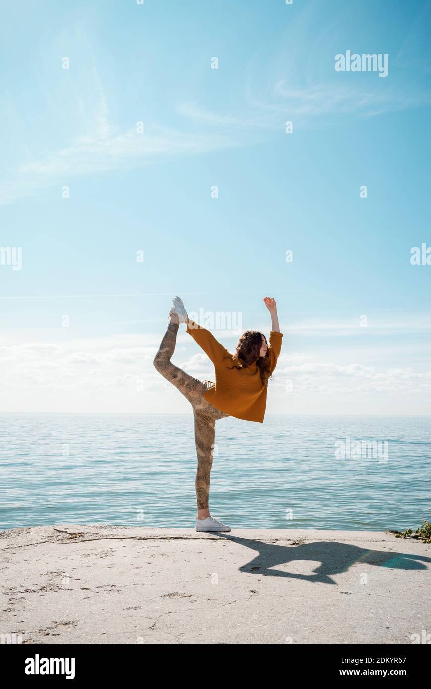 Giovane donna in un maglione su una spiaggia che pratica yoga Posa ballerina (Natarajasana) Foto Stock