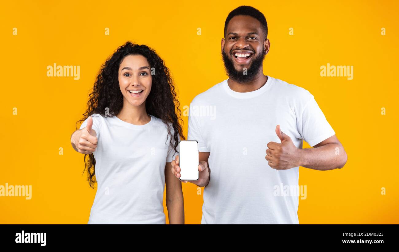 Coppia eccitata che mostra lo schermo del telefono gesturando Thumbs-Up su sfondo giallo Foto Stock