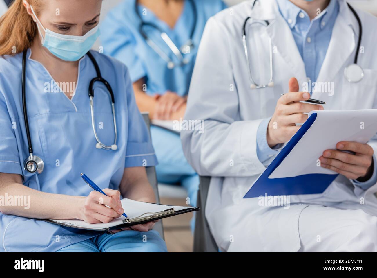 Infermiera che scrive sulla clipboard mentre si siede vicino al medico durante la riunione con colleghi offuscati in background in ospedale Foto Stock