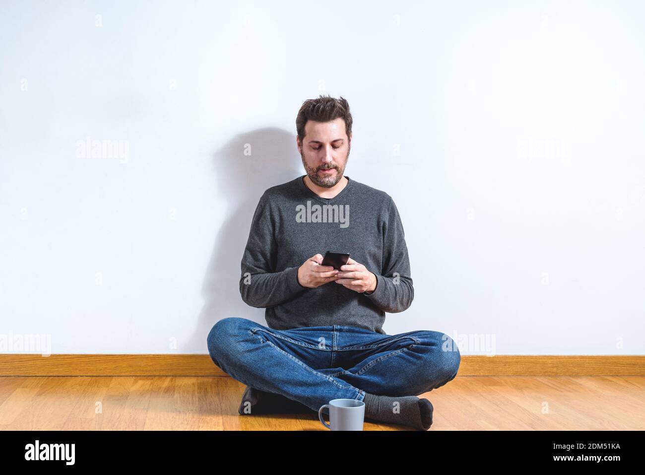 uomo caucasico sul suo vestito da 40 anni casual con un grigio maglione e jeans blu seduti con gambe incrociate sul pavimento in legno in un appartamento Foto Stock