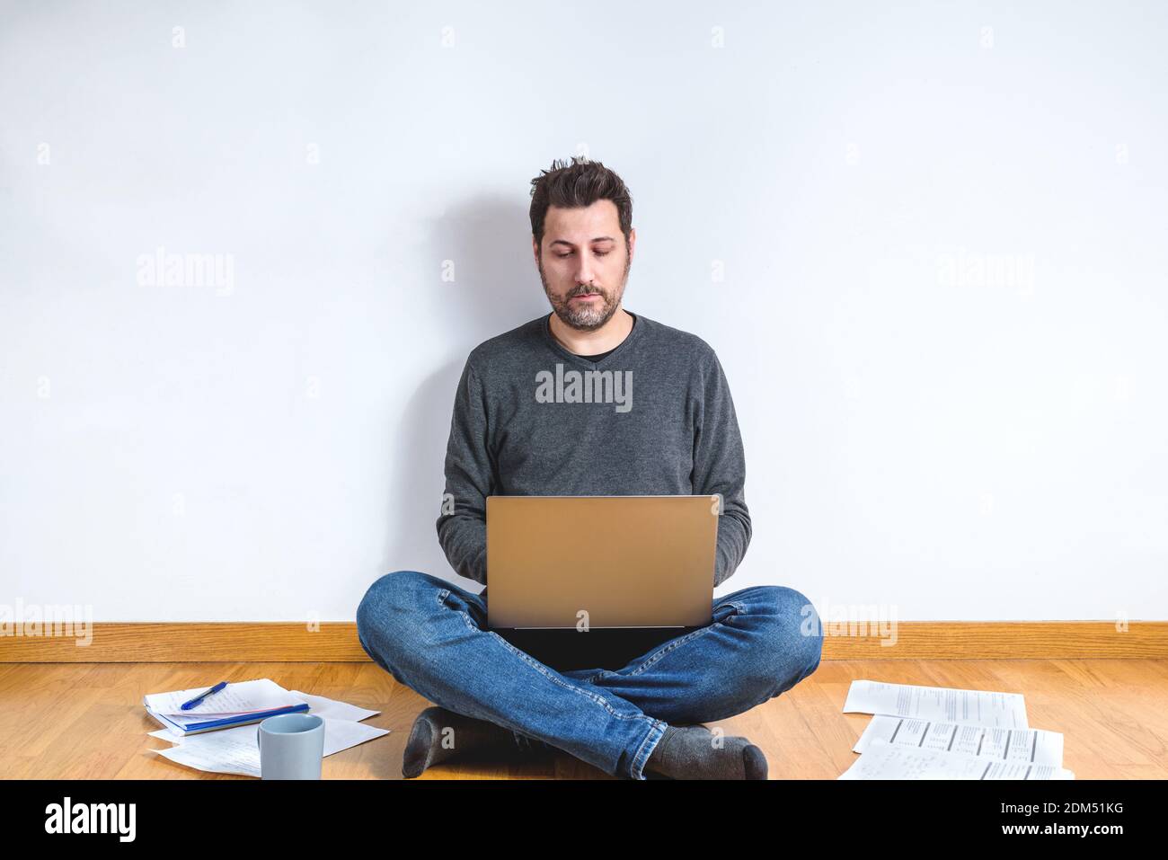 uomo caucasico sui suoi 40s seduto casual seduta sul pavimento in legno di un appartamento con un computer portatile e circondato di carte Foto Stock