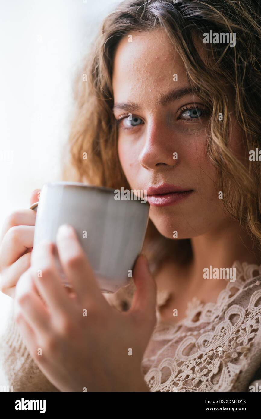 Giovane femmina con occhi blu sorseggiando una bevanda calda e guardando alla telecamera Foto Stock