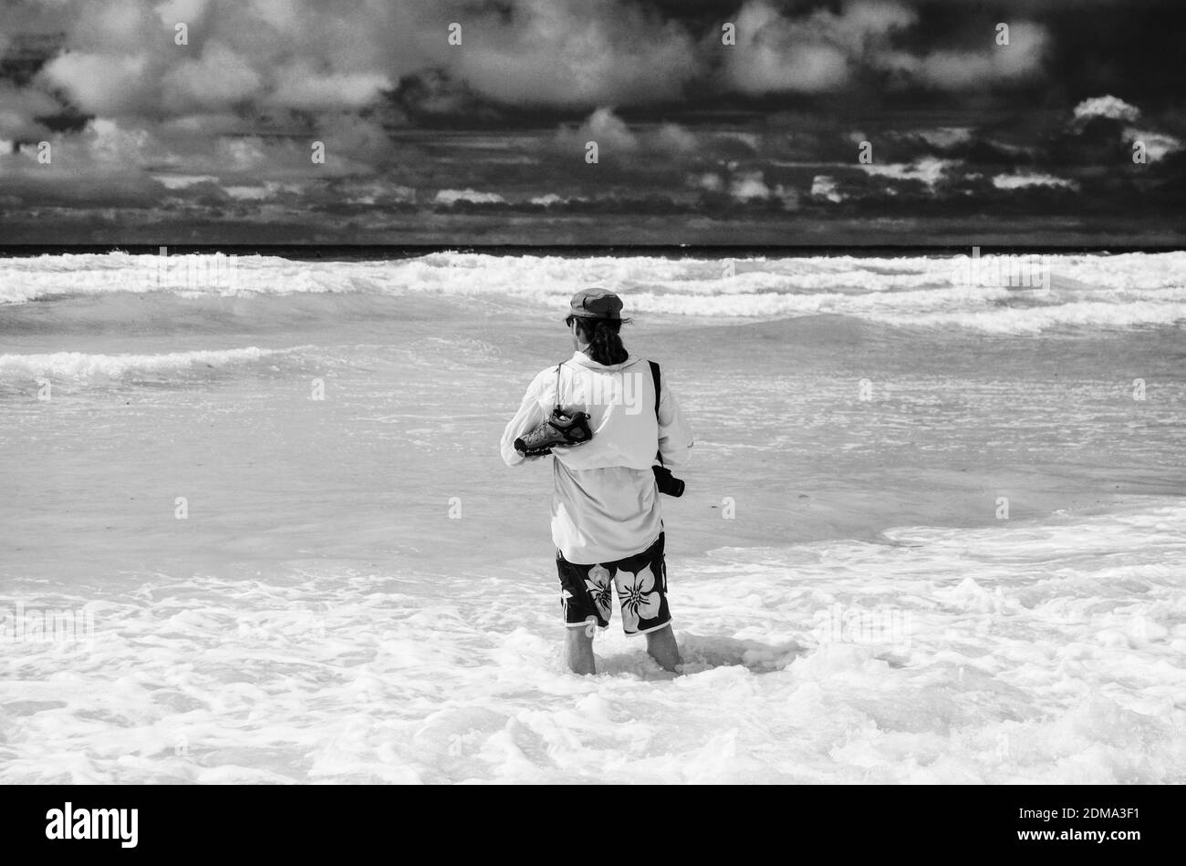 Uomo in beachwear in piedi ginocchio in profondità nell'oceano, sull'isola di Galapagos. Con macchina fotografica e scarpe sopra la spalla, con filtri in bianco e nero. Foto Stock