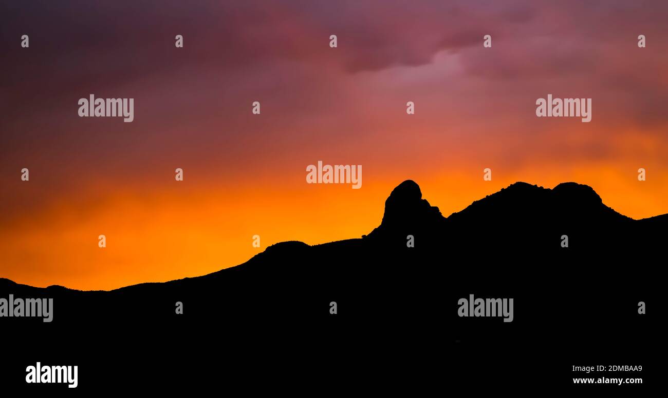 Tramonto attraverso le nuvole con raggi di luce. Viola e arancione vibranti con silhouette di montagna nera all'orizzonte. Foto Stock