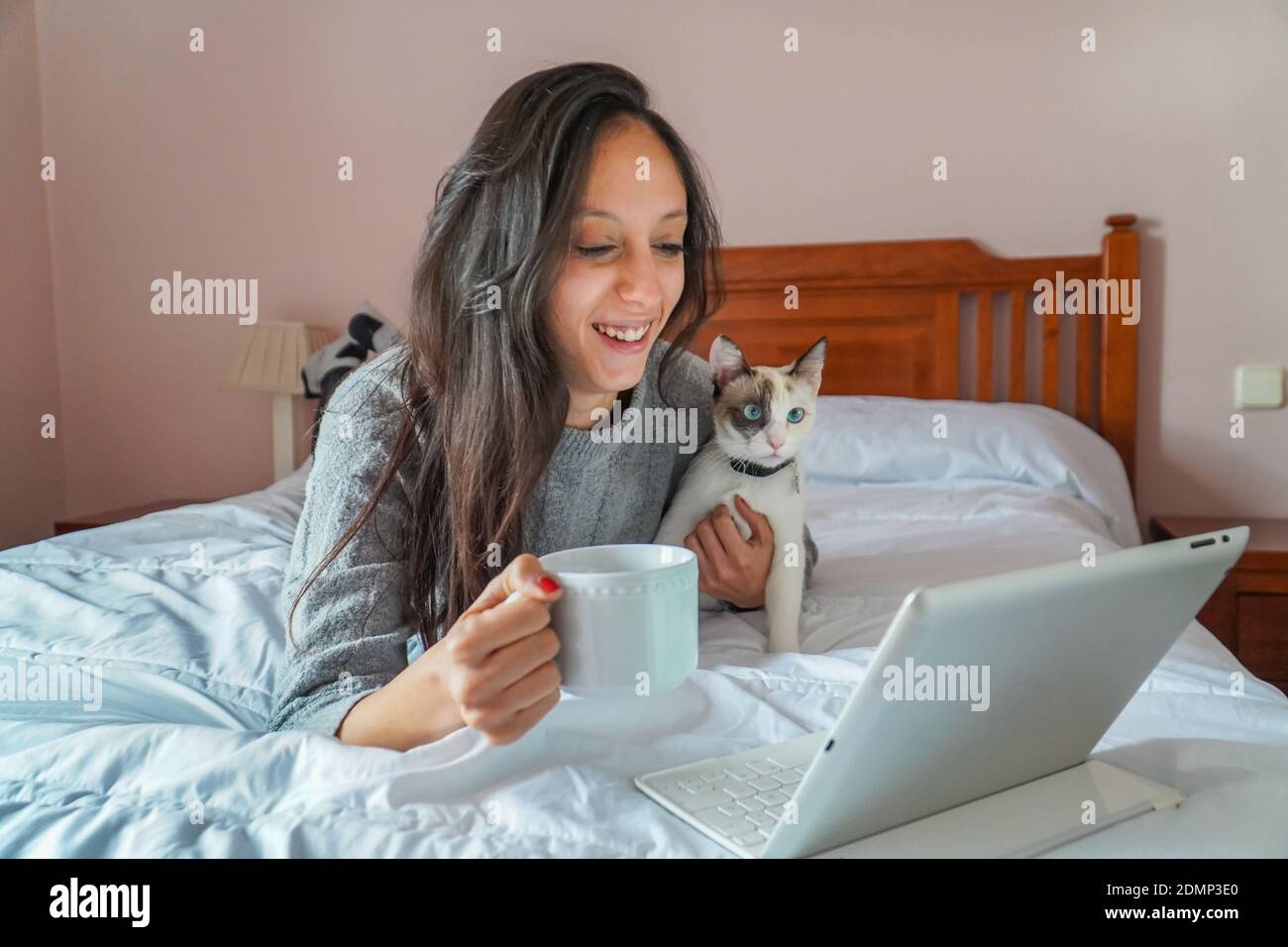 Una giovane donna in videoconferenza con il suo gatto e una tazza di caffè sul letto. Concetto quarentino e nuova normalità Foto Stock