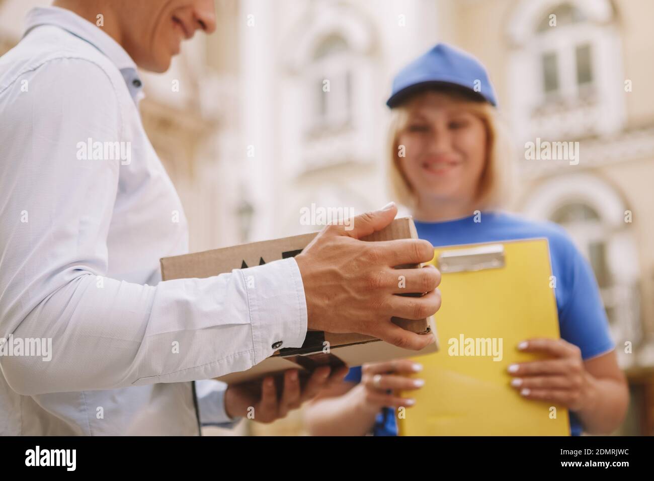 Primo piano corto di un uomo che tiene scatola di cartone, felice corrieri femmina sorridente sullo sfondo. Cliente soddisfatto sorridente, tenendo deliv. Scatola posta pacchi Foto Stock