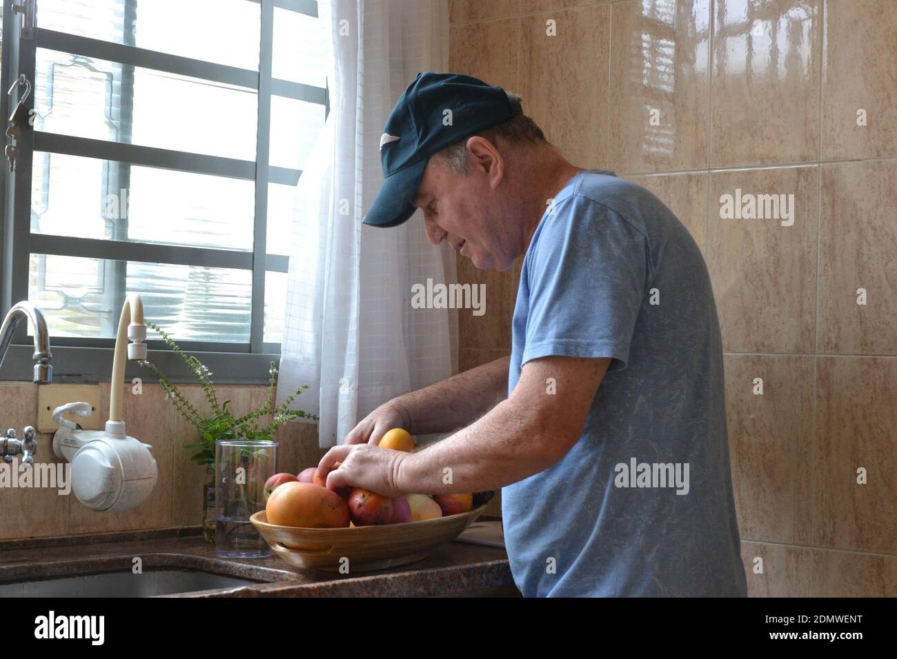 Uomo anziano uomo con cappello, scegliendo frutta per preparare il pasto di frutta a casa, con ciotola di frutta in legno, lavello e rubinetti, Brasile, Sud America Foto Stock
