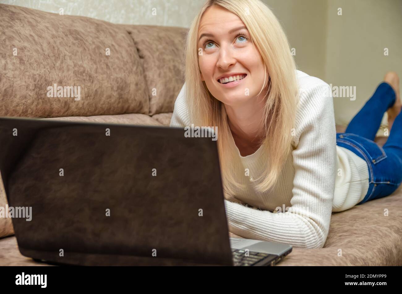 Young blonde woman is lying on the sofa with a laptop, smiling and looking up mysteriously. Concept of online communication, self-isolation Foto Stock