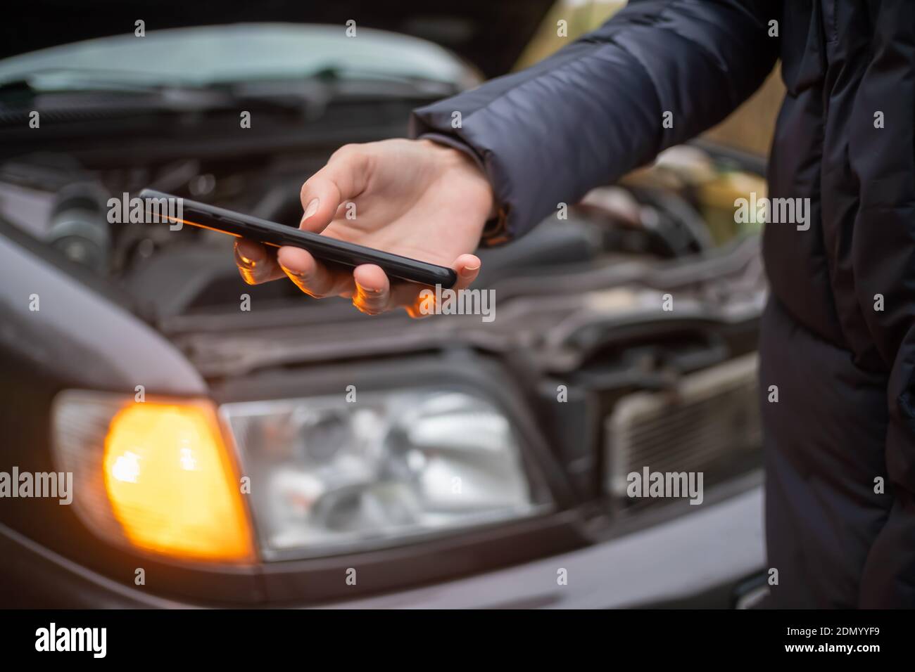 La mano della donna in una giacca tiene un telefono cellulare nero primo piano contro il cofano aperto di un'auto su un arresto di emergenza Foto Stock