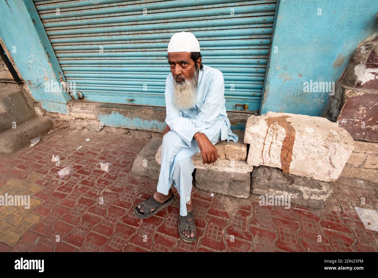 Jamnagar, Gujarat, India - Dicembre 2018: Un ritratto di un anziano indiano con una barba che indossa un cappello bianco del cranio seduto sul marciapiede. Foto Stock