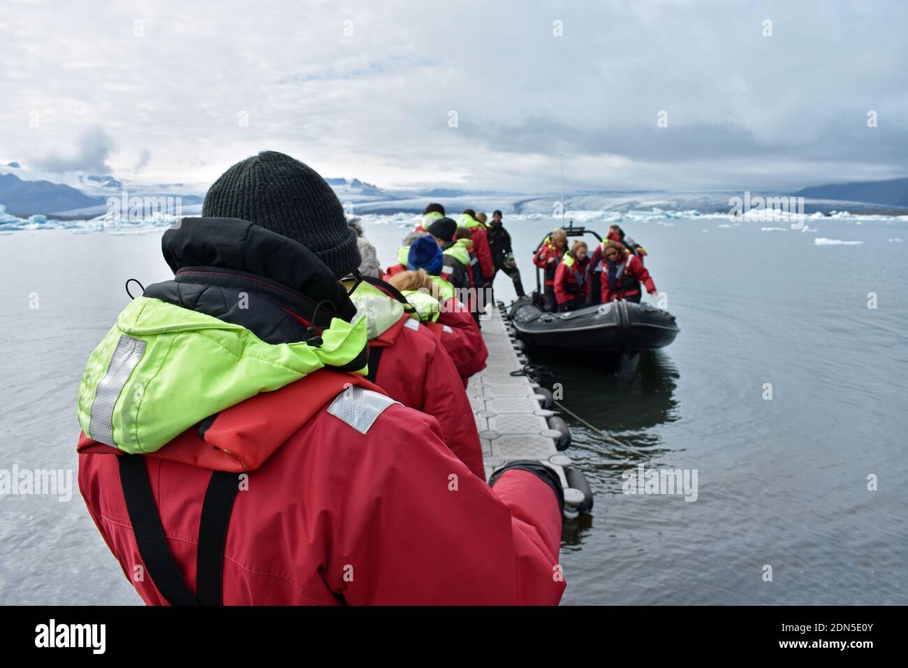 Un gruppo di tour vestito di cappotti rossi e giubbotti salvagente si allinea su un pontile galleggiante per salire a bordo delle barche zodiacali nella laguna glaciale di Jokulsarlon, Islanda del Sud. Foto Stock