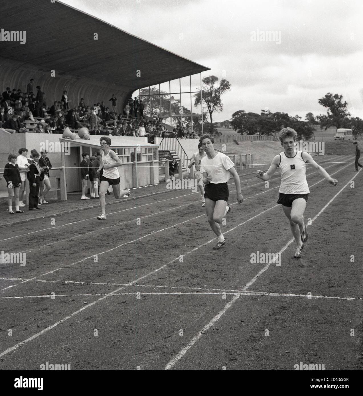 1960s, storico, guardato dagli spettatori seduti in una tribuna, i giovani che gareggiano in una corsa fuori su una pista di cenere, in una giornata sportiva della contea, Fife, Scozia, Foto Stock