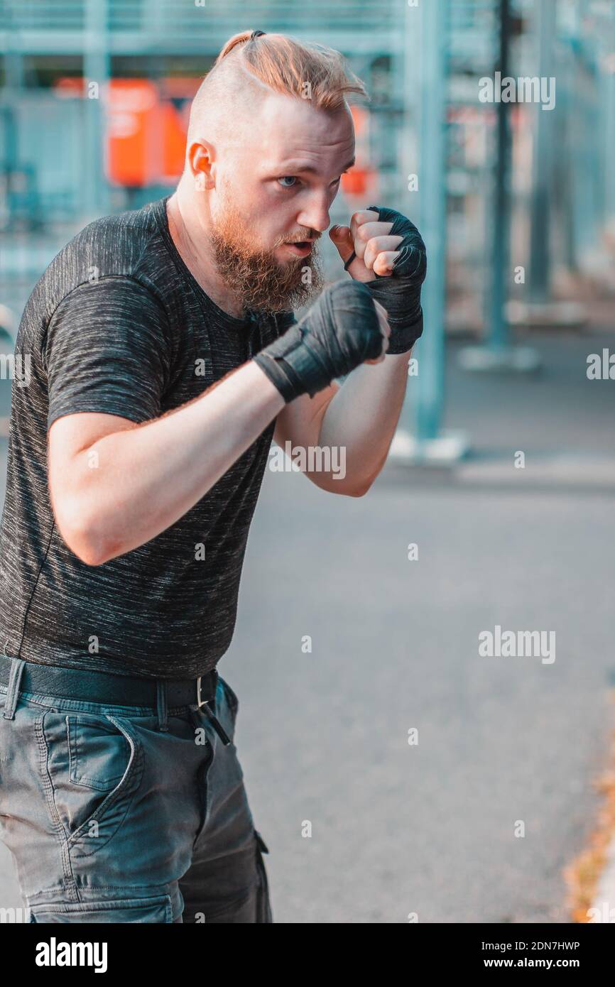 Street Fighter in vestiti neri e bende sulla boxe del polso in sacchetto di punzonatura all'aperto. Young Man facendo Box Training e praticando i suoi pugni alla palestra esterna Foto Stock