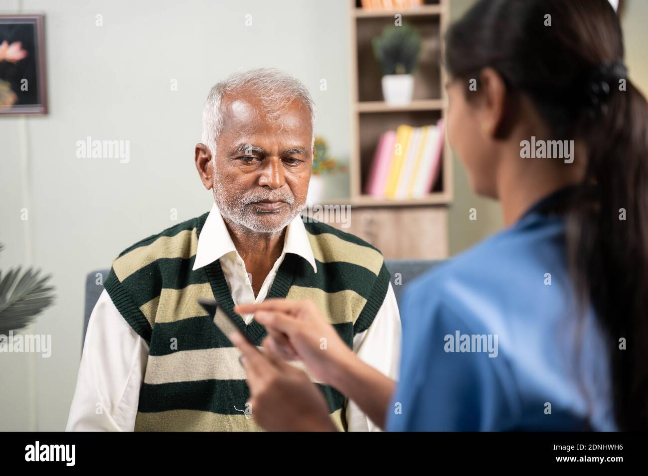 Colpo di spalla del medico che spiega al paziente i dettagli sulla salute utilizzando l'app mobile sul telefono durante la visita di routine a casa. Foto Stock