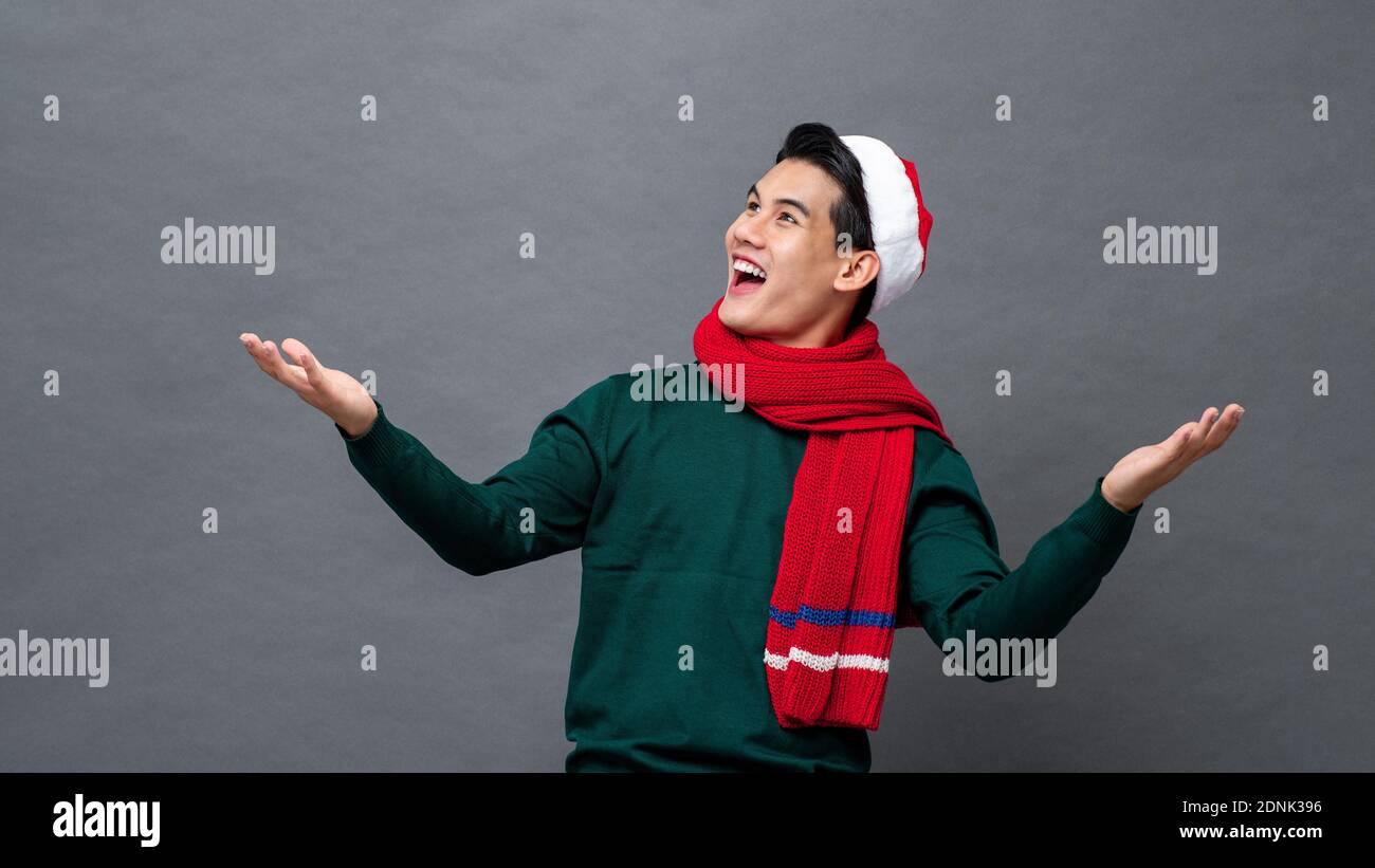 Giovane uomo asiatico divertito che indossa abiti a tema natalizio con aperto gesto palmo che cerca di svuotare lo spazio da parte Foto Stock