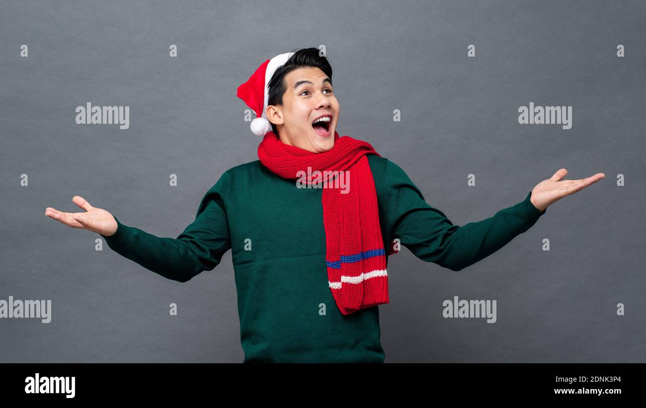 Giovane uomo asiatico divertito che indossa abiti a tema natalizio con aperto gesto palmo che cerca di svuotare lo spazio da parte Foto Stock