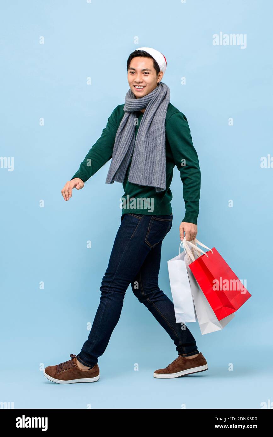 Handsosme uomo asiatico in tema di Natale attrie a piedi e tenere borse per lo shopping in uno studio di colore azzurro Foto Stock