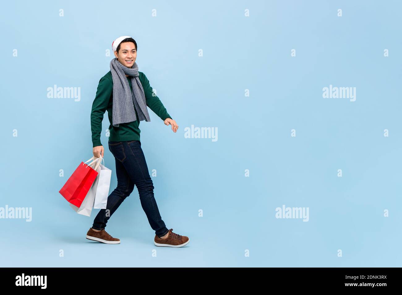 Handsosme uomo asiatico in tema di Natale attrie a piedi e tenere borse per lo shopping in uno spazio da studio blu chiaro con spazio per la copia Foto Stock