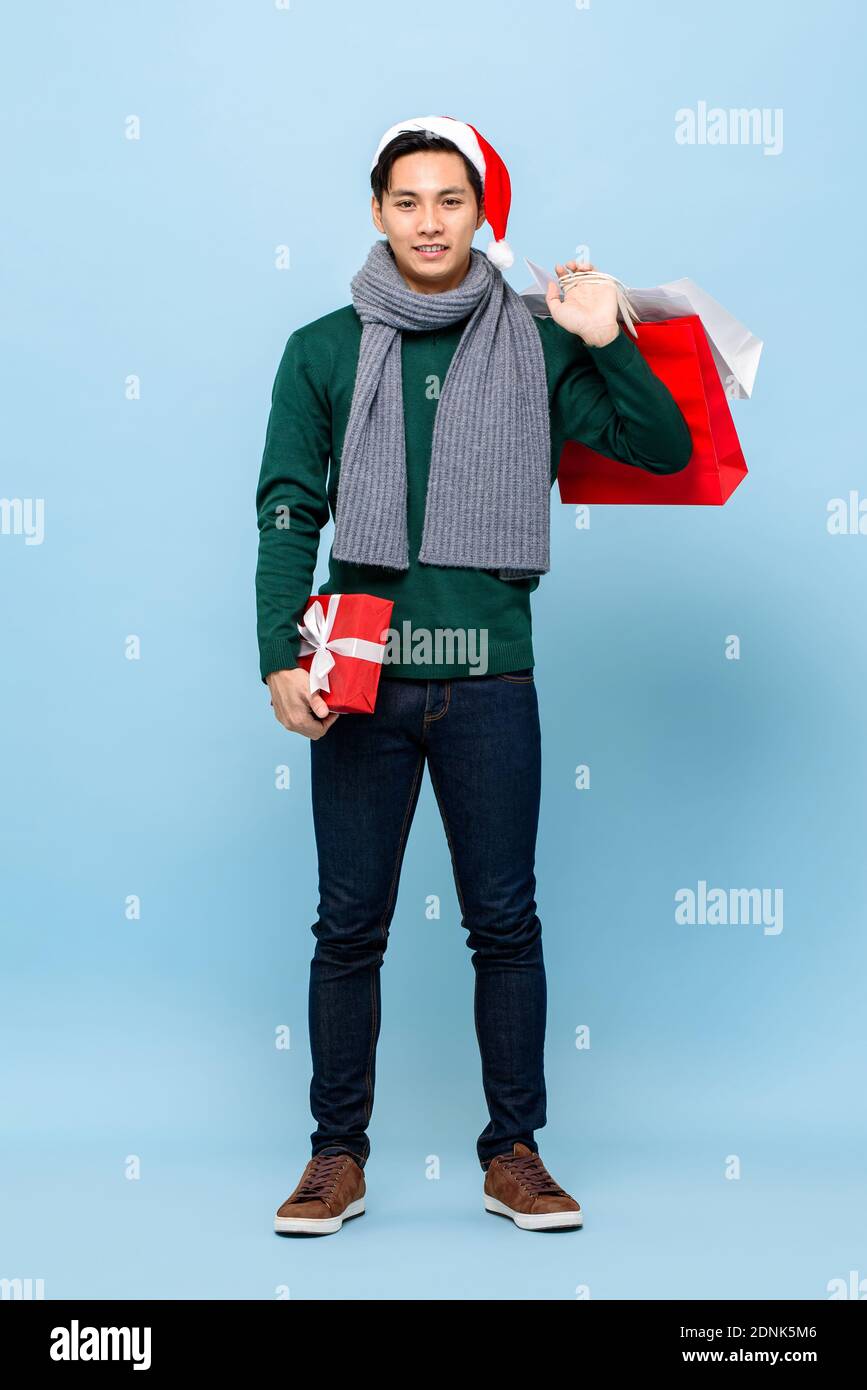 Ritratto a lunghezza intera di sorridente felice giovane bell'uomo asiatico indossa un pullover di natale verde e un cappello rosso che contiene una confezione regalo e borse per la spesa isolati Foto Stock
