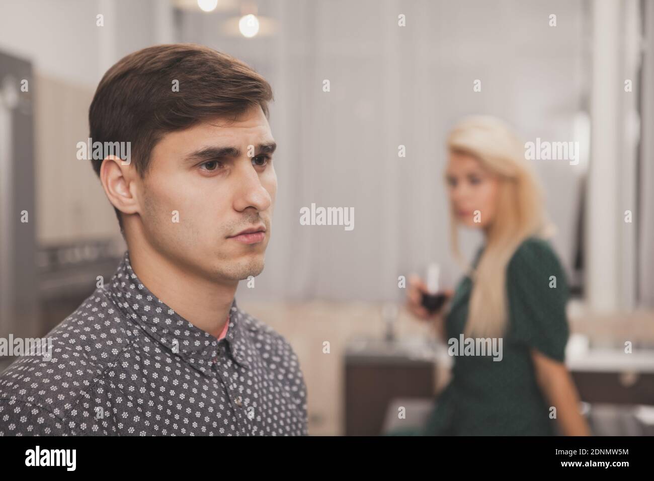 Bell'uomo che guarda via pensieroso, sua moglie beve vino sullo sfondo. Uomo depresso che guarda sconvolto dopo aver litigato con sua moglie. Coppia f Foto Stock