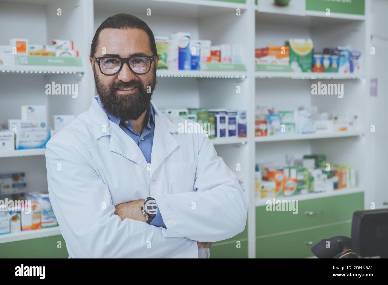 Proud mature male pharmacist smiling to the camera with arms crossed, posing at his new local drugstore. Cheerful chemist welcoming customers in his p Foto Stock