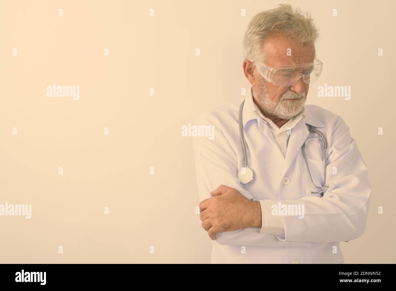 Studio shot di sad senior uomo barbuto medico pensando indossando occhiali protettivi con le braccia incrociate contro uno sfondo bianco Foto Stock