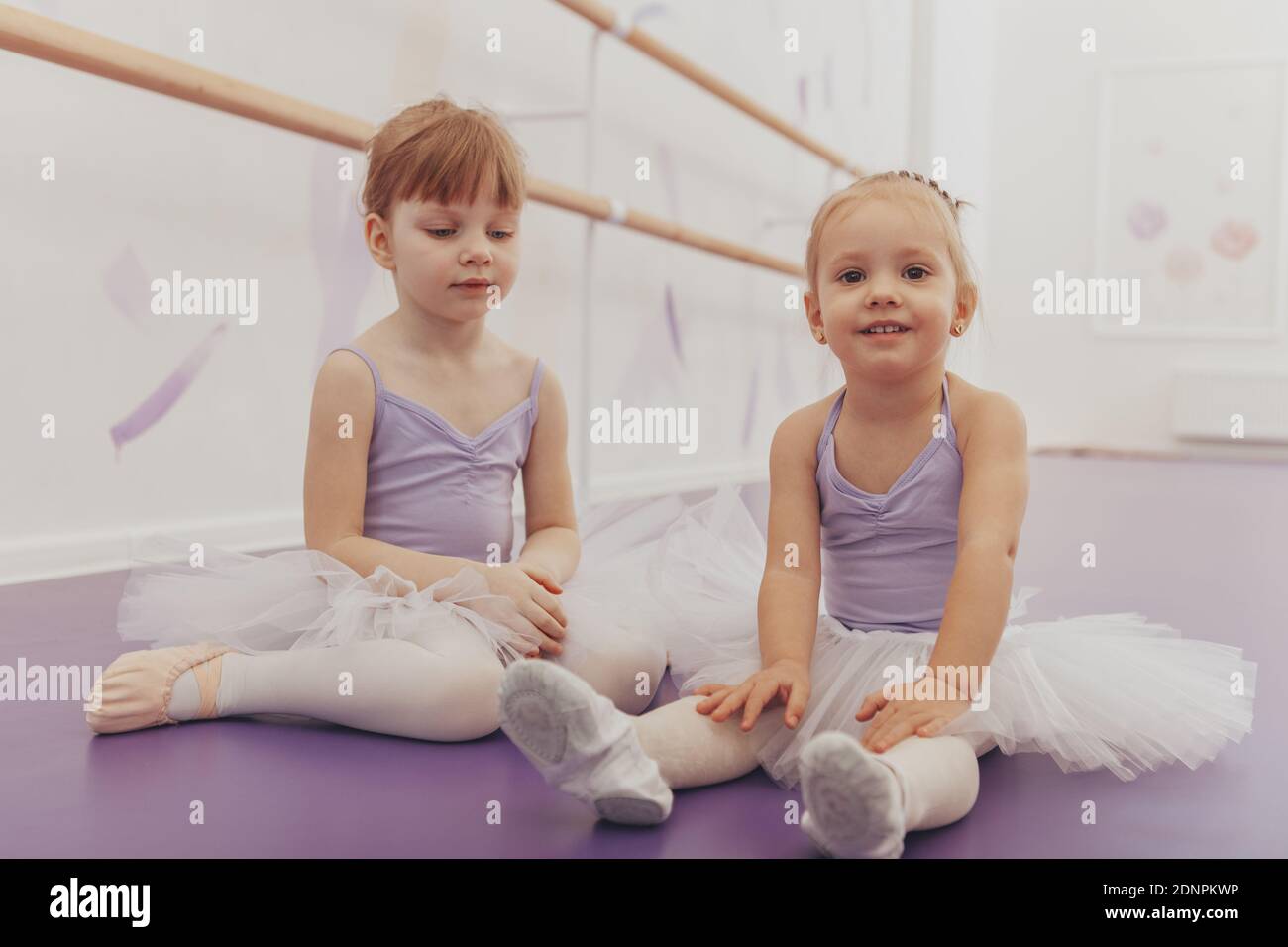 Due adorabili ballerine allegre riposano sul pavimento presso la sala da ballo. Carina ragazza felice in abito da ballo sorridente gioiosamente, seduta con lei Foto Stock