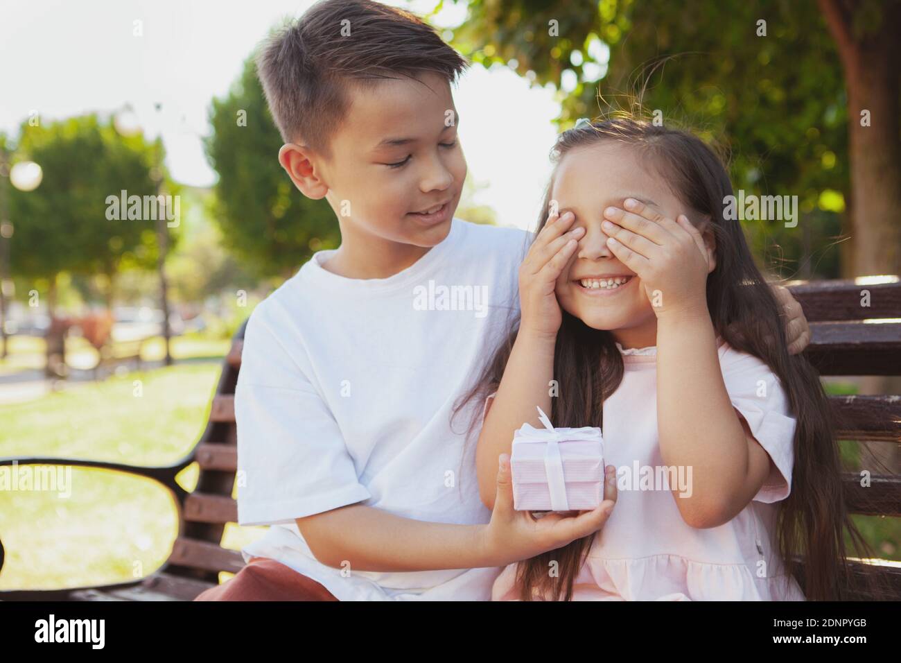 Allegro ragazzo asiatico che dà un regalo a sorpresa alla sua sorellina. Adorabile bambina che le copre gli occhi con le mani, ricevendo un regalo da Foto Stock