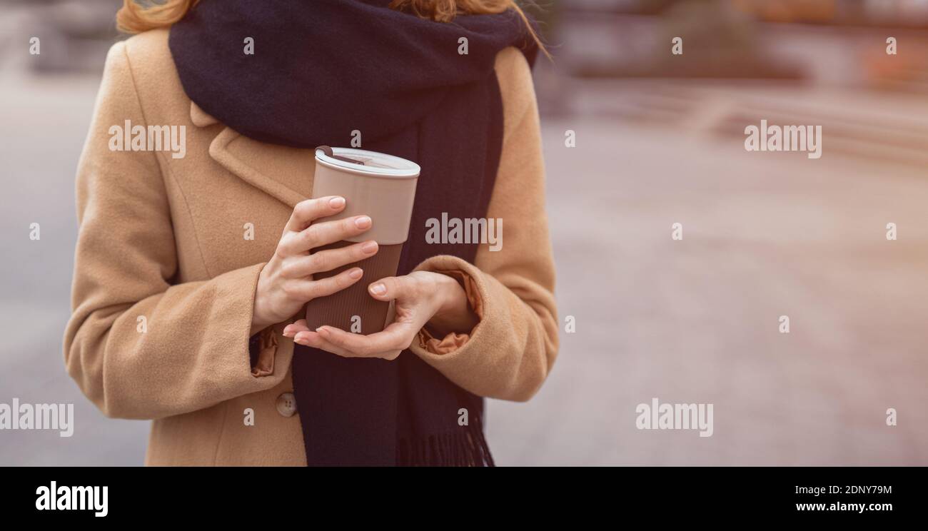 Ritratto delle mani di una donna che reggono una tazza da caffè con cappotto beige e sciarpa nera in piedi all'aperto. Elegante giovane donna con outfit autunnale Foto Stock