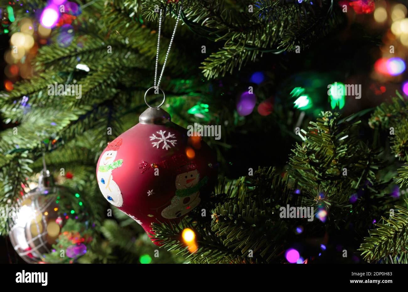 primo piano di un ornamento rosso dell'albero di natale con un pupazzo di neve appeso ad un albero di natale con luci bokeh scintillanti intorno Foto Stock