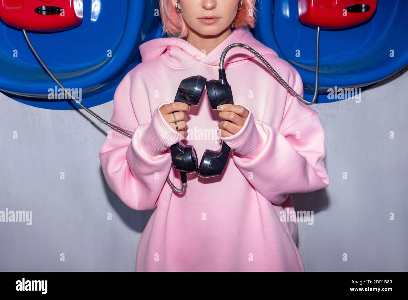 Giovane donna che indossa una camicia con cappuccio rosa davanti alle cabine telefoniche, che tiene i ricevitori Foto Stock