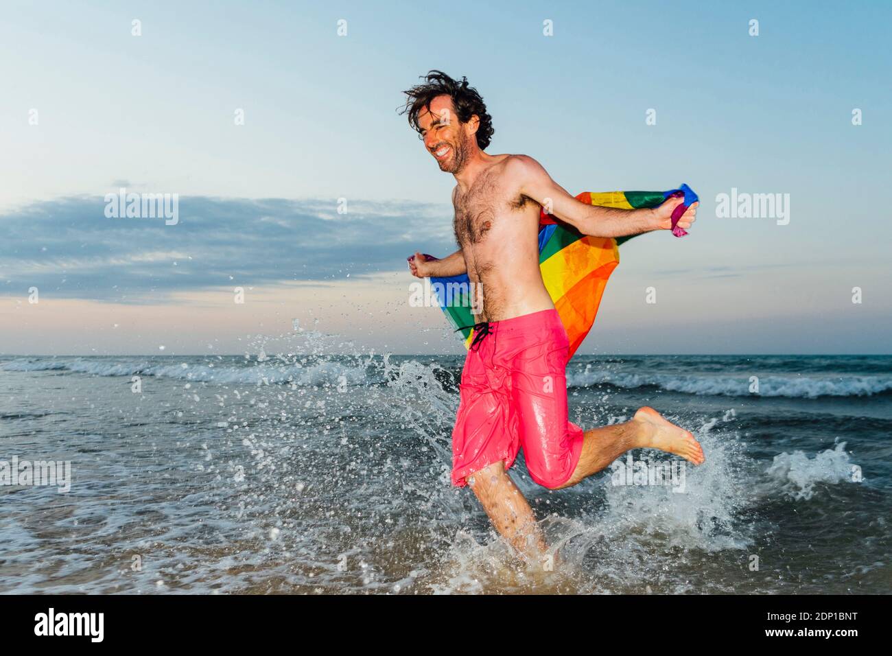 Ridendo uomo che corre in acqua, portando bandiera gay orgoglio Foto Stock