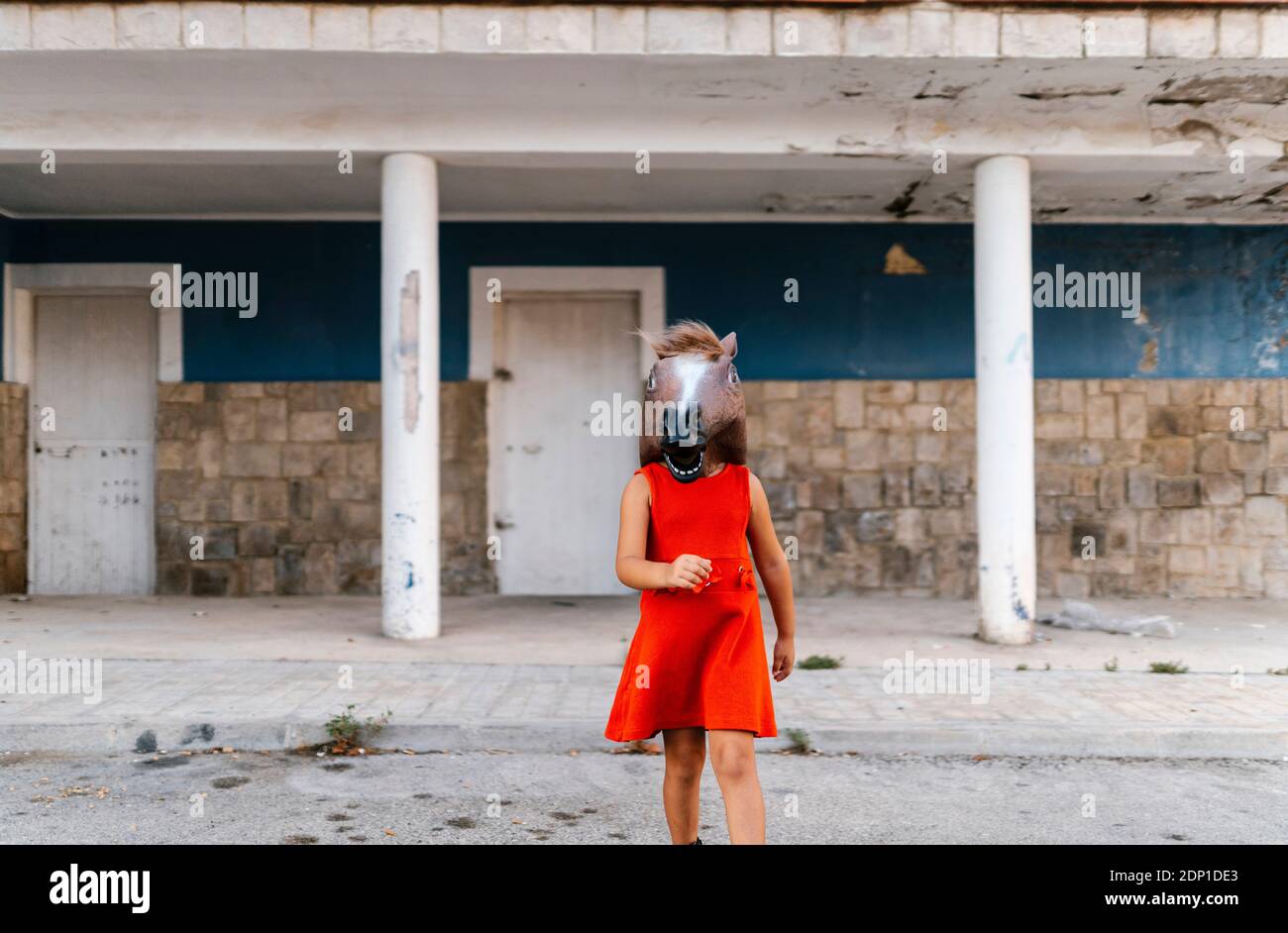 Bambina con testa di cavallo e vestito rosso a piedi da una casa abbandonata Foto Stock