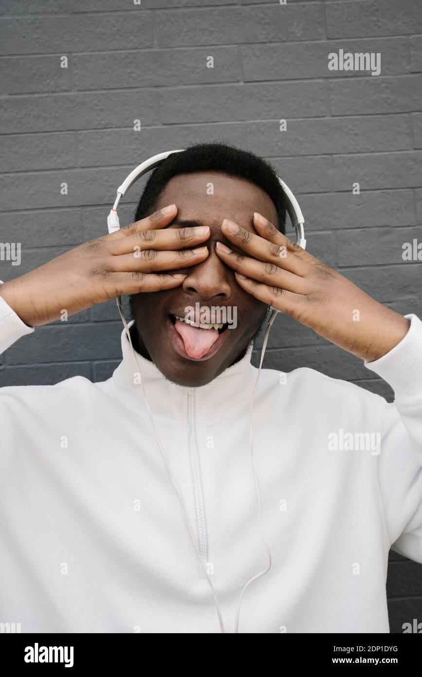 Ritratto di un giovane che ascolta la musica con le cuffie che coprono gli occhi durante l'estrazione della lingua Foto Stock